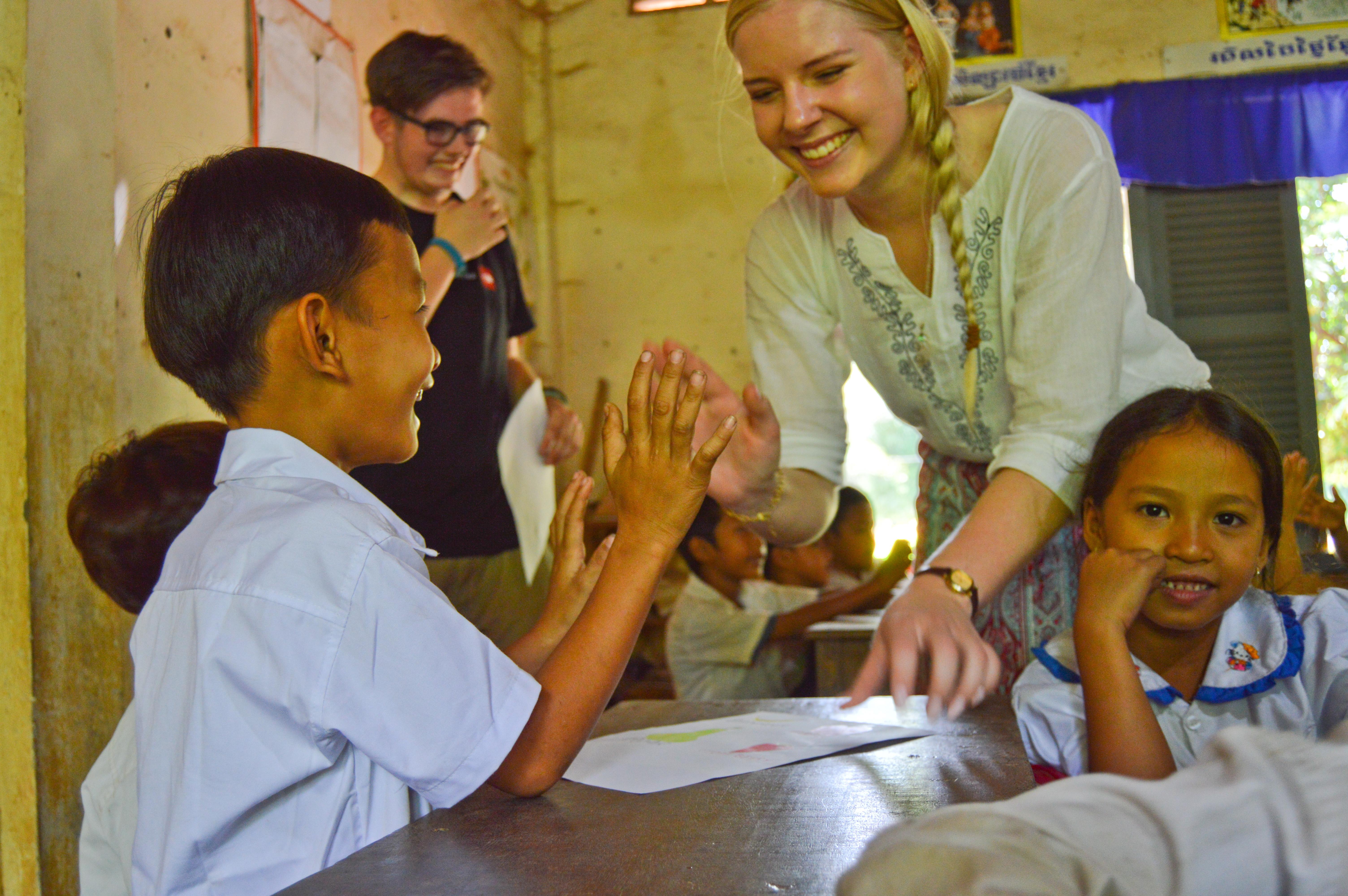 As part of the Projects Abroad community teenage work in Cambodia, volunteers help teachers in the classroom with work. A teenage volunteer working with children in Cambodia gives a young child a high-five in the classroom.